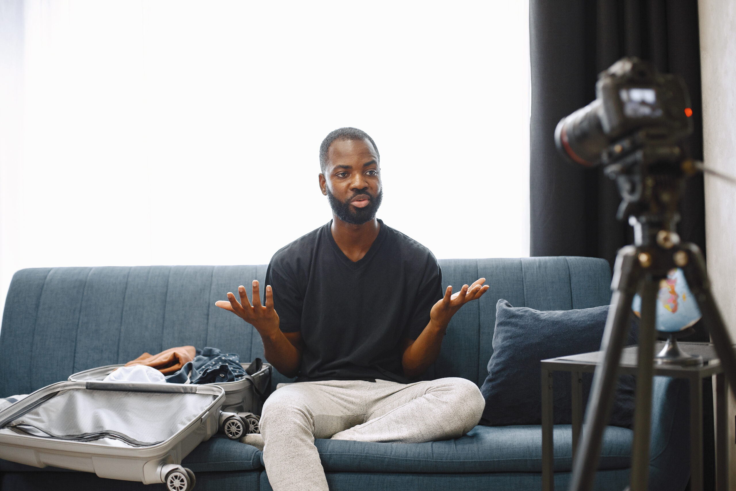 African male blogger recording vlog on digital camera African-american male blogger sitting in front of camera and recording a video about his luggage. Man wearing dark t-shirt and white trousers. Blogger speaking about traveling.