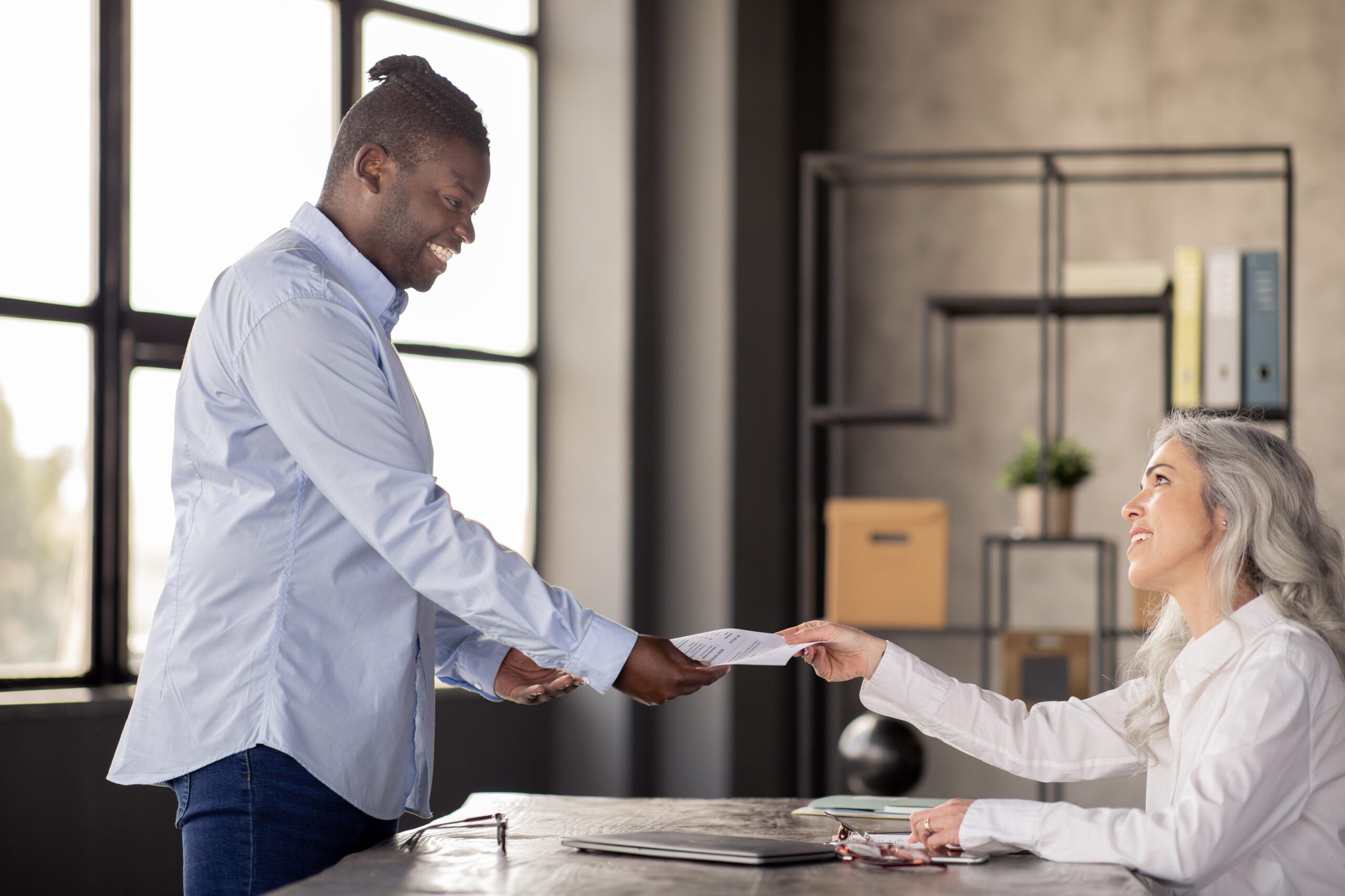African Man Giving Resume To HR Manager Woman In Office Employment Offer. African American Man Giving His Paper CV Resume To HR Manager Woman, Applying For Job Vacancy During Job Interview In Office Indoors. Recruitment And Human Resources Concept